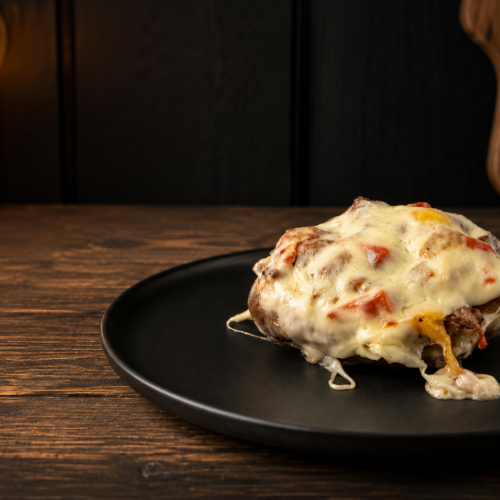 Philly cheesesteak stuffed portobello mushroom topped with melted cheese and peppers on a black plate over a rustic wood table.